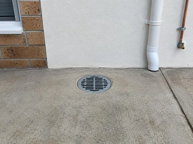 Outdoor gully grate set in dry concrete paving beside a house wall in Penrith