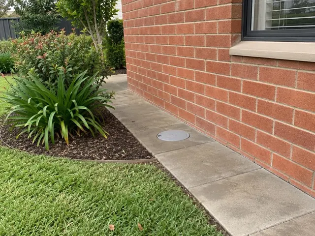Concrete path beside red brick wall with a circular capped access point and tidy garden in Cambridge Park