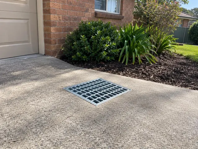 Concrete driveway with a flush stormwater grate beside a brick house and garden in Kingswood