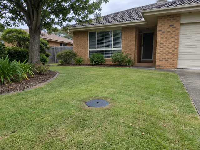 Front lawn of a brick house with a round capped sewer opening in the grass in Glenmore Park